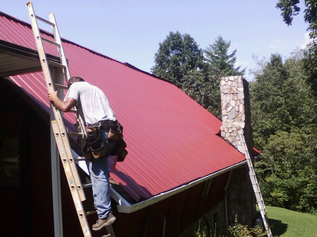 Roofer on a ladder beside a red metal roof and stone chimney — Soffit & Fascia Repair by Roofing Brothers in Joppa